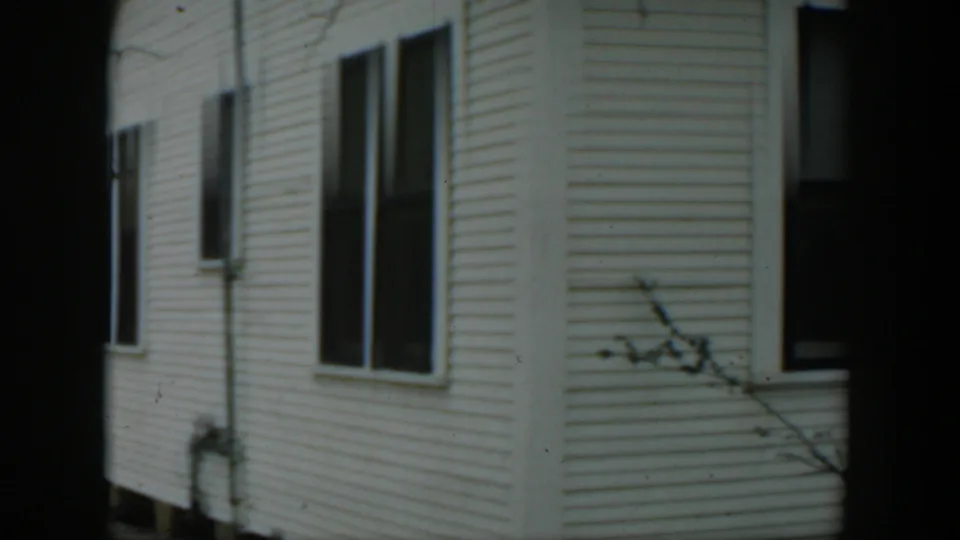 1951 Cuba 8mm frame of a white clapboard house corner, dark windows, exterior piping, and muted overcast light.