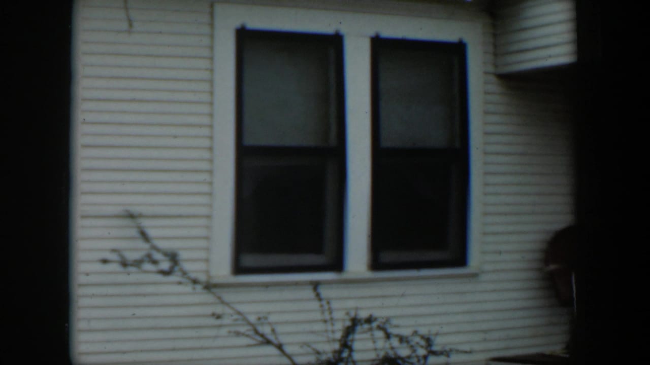 1951 Cuba home-movie frame showing a white clapboard house wall, two dark windows, and thin branches in the foreground.