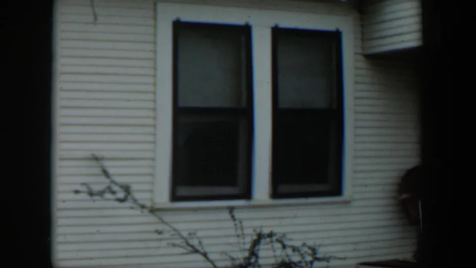 1951 Cuba home-movie frame showing a white clapboard house wall, two dark windows, and thin branches in the foreground.