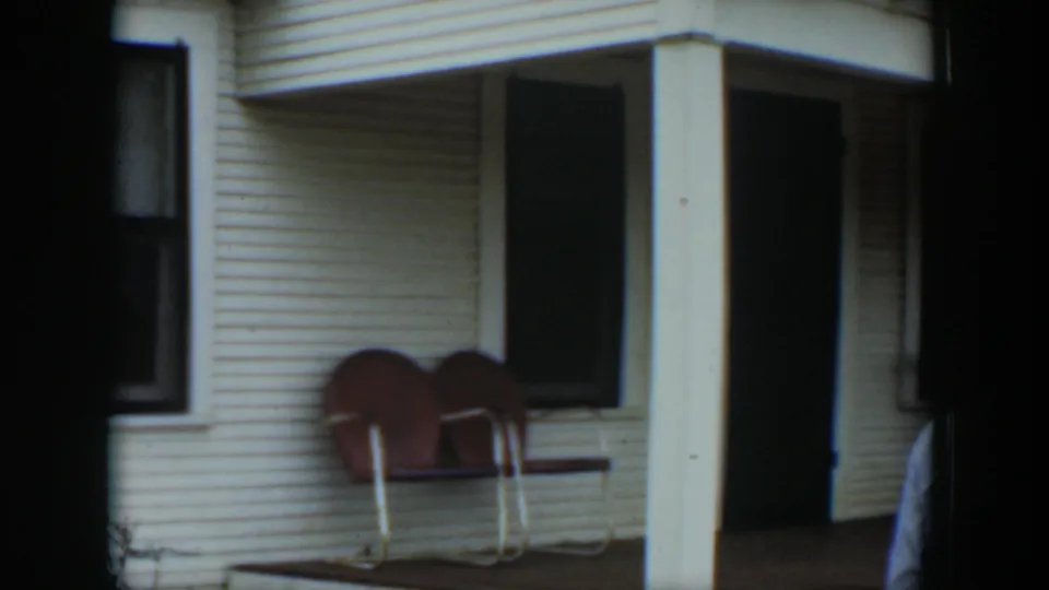 1951 Cuba frame with two brown chairs against a white clapboard porch, a square support post, and a dark doorway.
