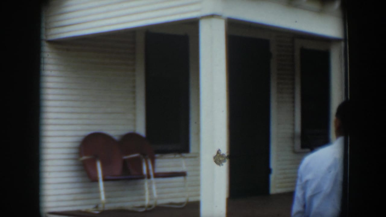 In Cuba home-movie footage from 1951, a white clapboard porch with red chairs is seen as a man in a light shirt enters the frame.