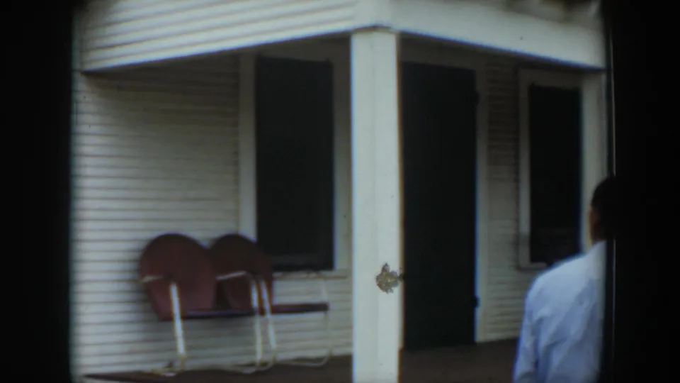 In Cuba home-movie footage from 1951, a white clapboard porch with red chairs is seen as a man in a light shirt enters the frame.