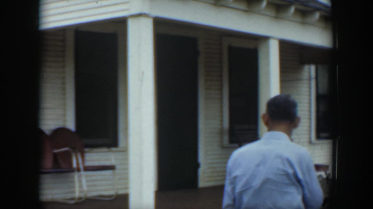 Man in a light shirt walks toward a shaded porch and dark doorway.