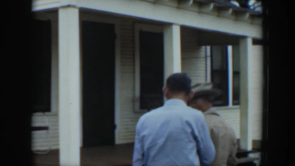Two adult men appear near the front porch of a white clapboard house, one seen from behind and another wearing a brimmed hat.