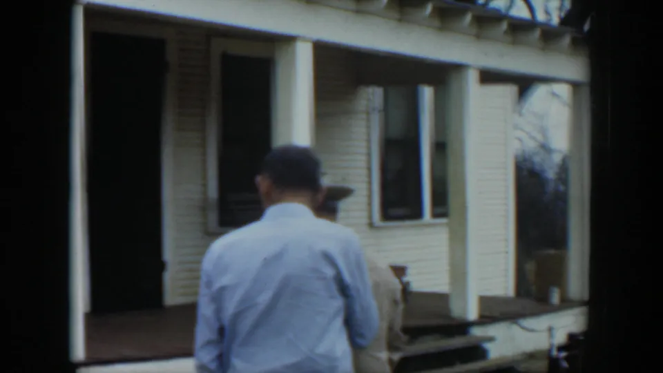 The man in the light shirt stands near the house porch with another adult partly visible at right in the Cuba 1951 home-movie scene.