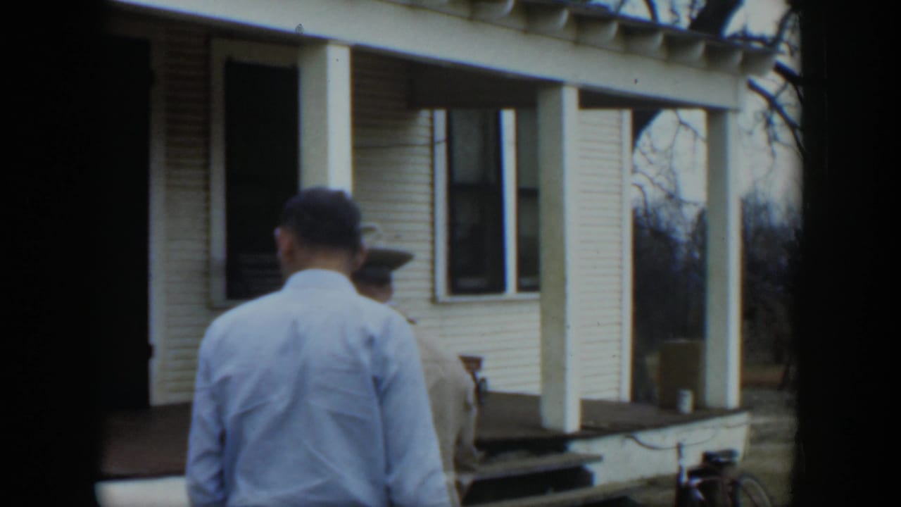 In 1951 Cuba footage, two men face a white clapboard house porch while a bicycle rests near the steps.