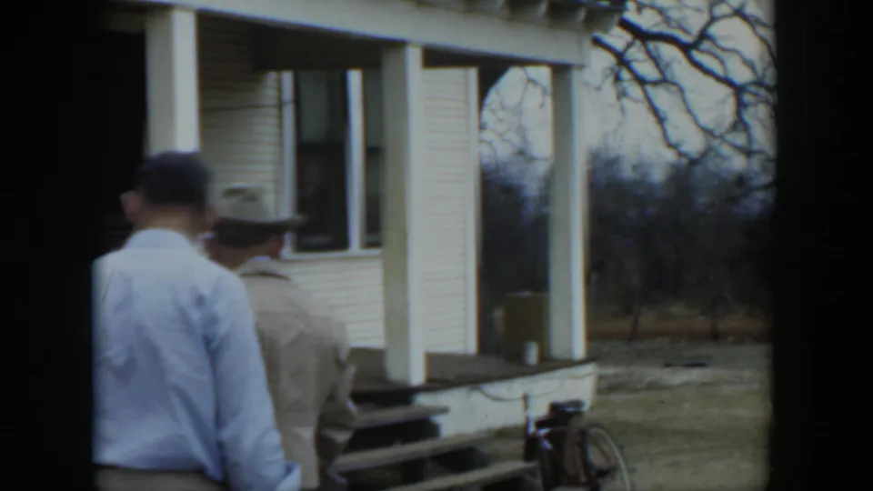 Two men in light clothing are seen from behind near the porch of a rural Cuban house, with a bicycle at the side.