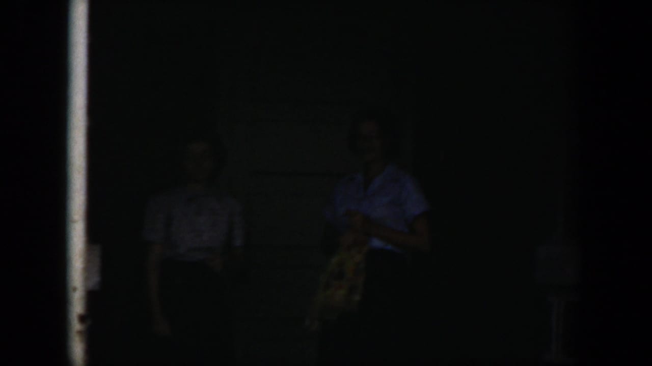 Two faint adult figures stand in deep shadow near a doorway.