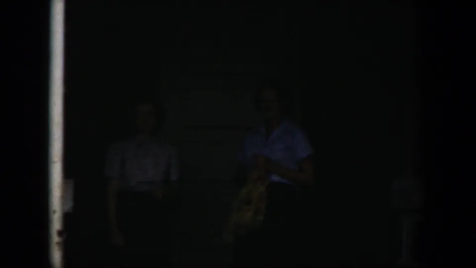 Two adult women appear faintly in the dark doorway area, with clothing and faces only partly visible in low light.