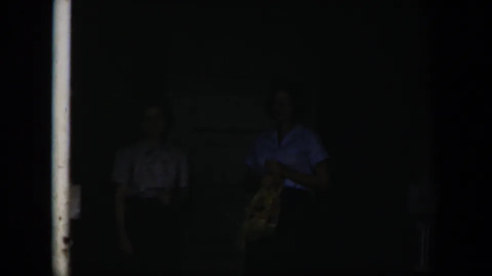 Two adult women stand near the doorway in dim light, with one woman holding a patterned bag and both figures partly obscured.