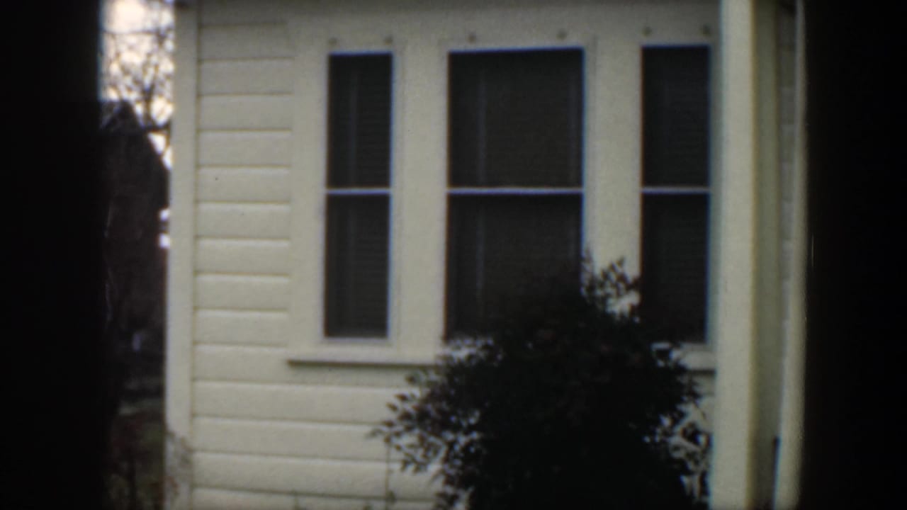 1951 Cuba home-movie frame showing a light-colored clapboard house facade with tall windows, porch details, shrubs, and heavy dark edge shadows.
