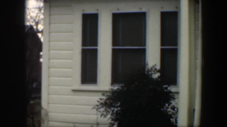 1951 Cuba home-movie frame showing a light-colored clapboard house facade with tall windows, porch details, shrubs, and heavy dark edge shadows.