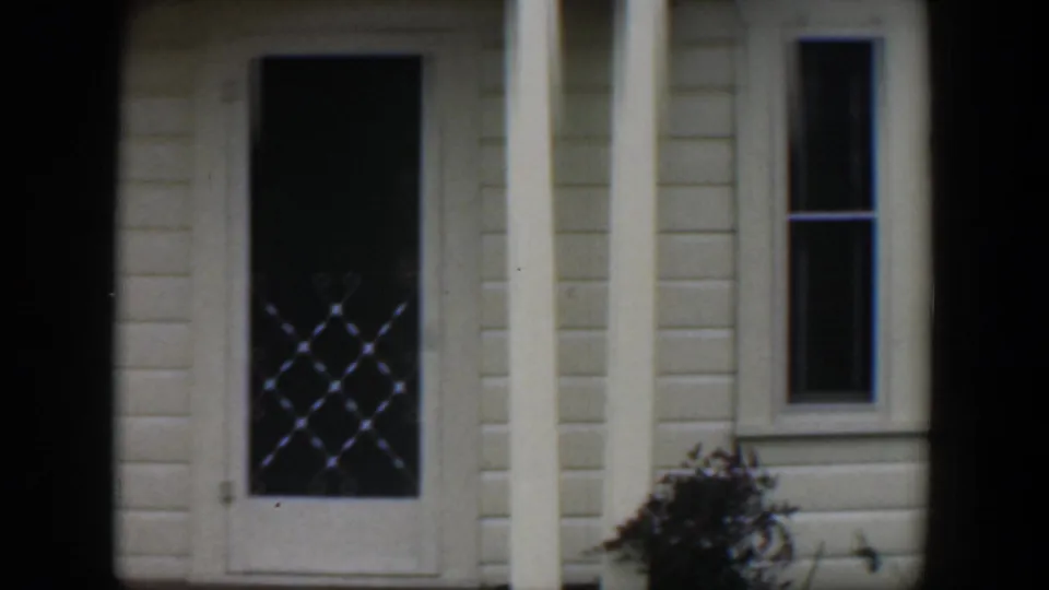 1951 Cuba home-movie frame showing the house entrance, including a door with decorative glass, porch post, narrow window, siding, and a small shrub.