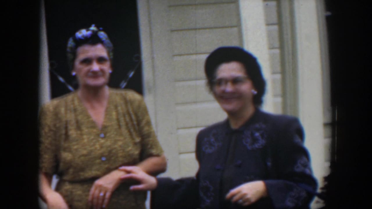 Two women standing outside a white house, one in a gold dress and headscarf, the other in a dark jacket and hat.
