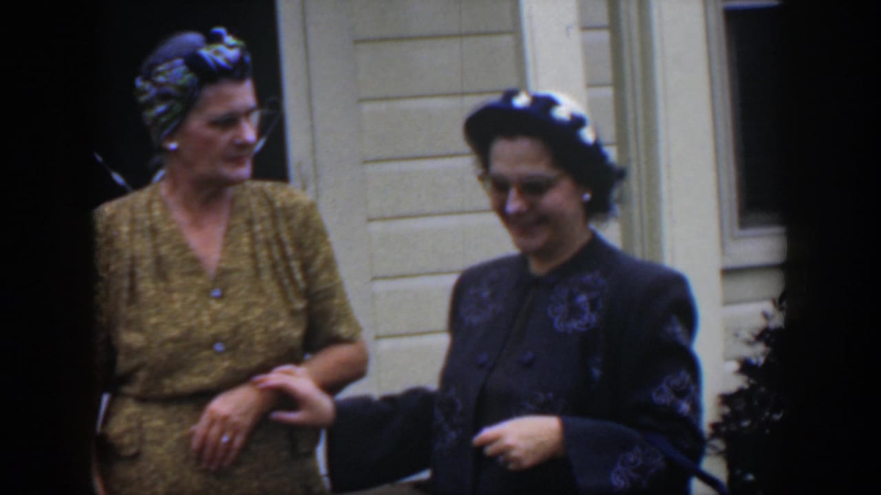 Two women outside a white house, with one woman looking toward the other near a doorway and window.