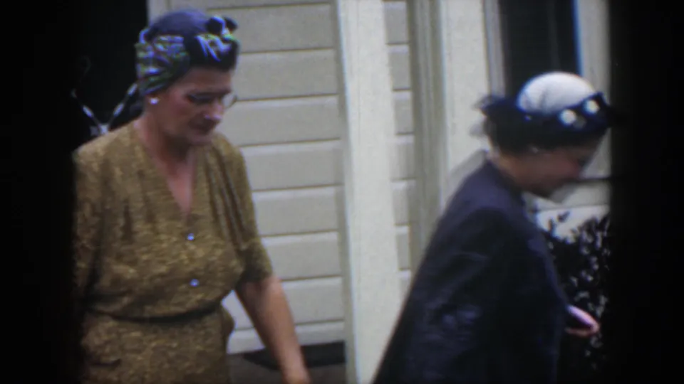 Two adult women walk beside a white house in Cuba, with visible glasses, a patterned headscarf, a gold dress, and a white-and-blue hat.