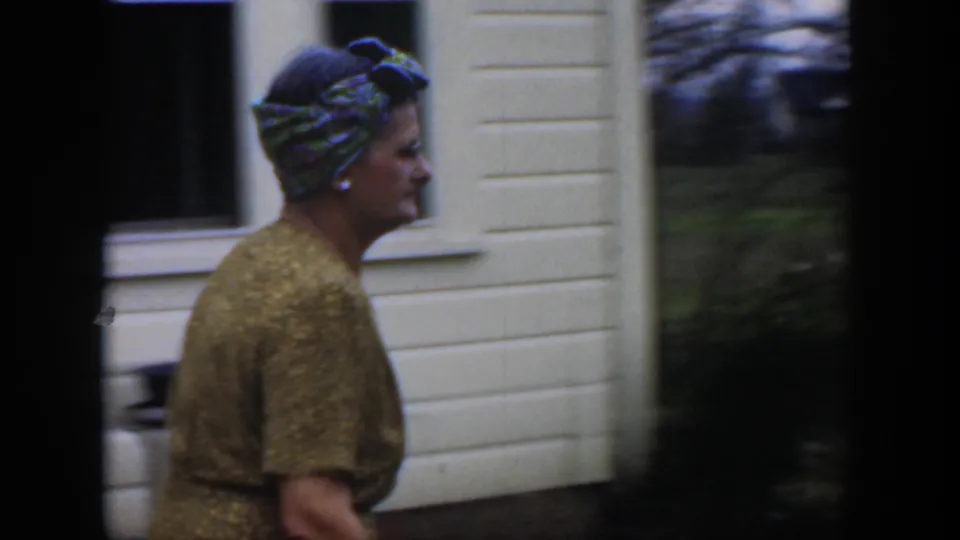 A woman in a gold-colored dress and patterned headscarf walks past a white house in 1951 Cuba home-movie footage.