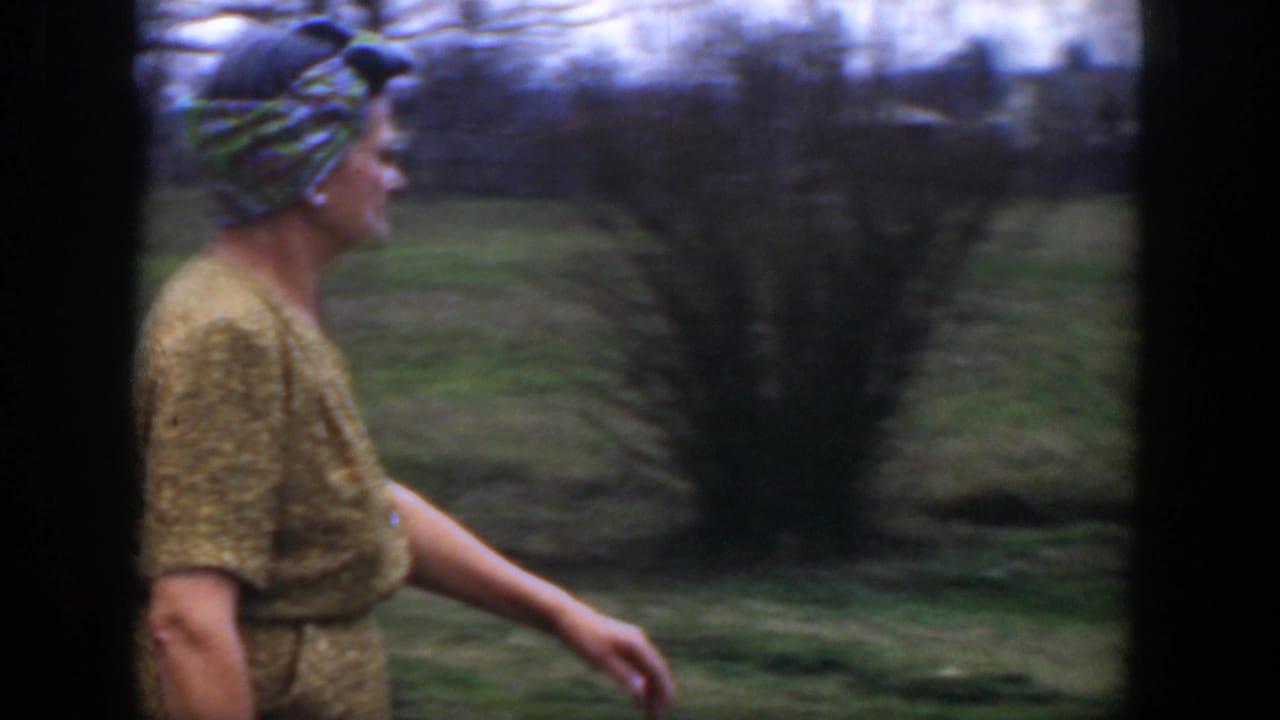 Woman in a yellow patterned dress and headscarf walks across a grassy yard.