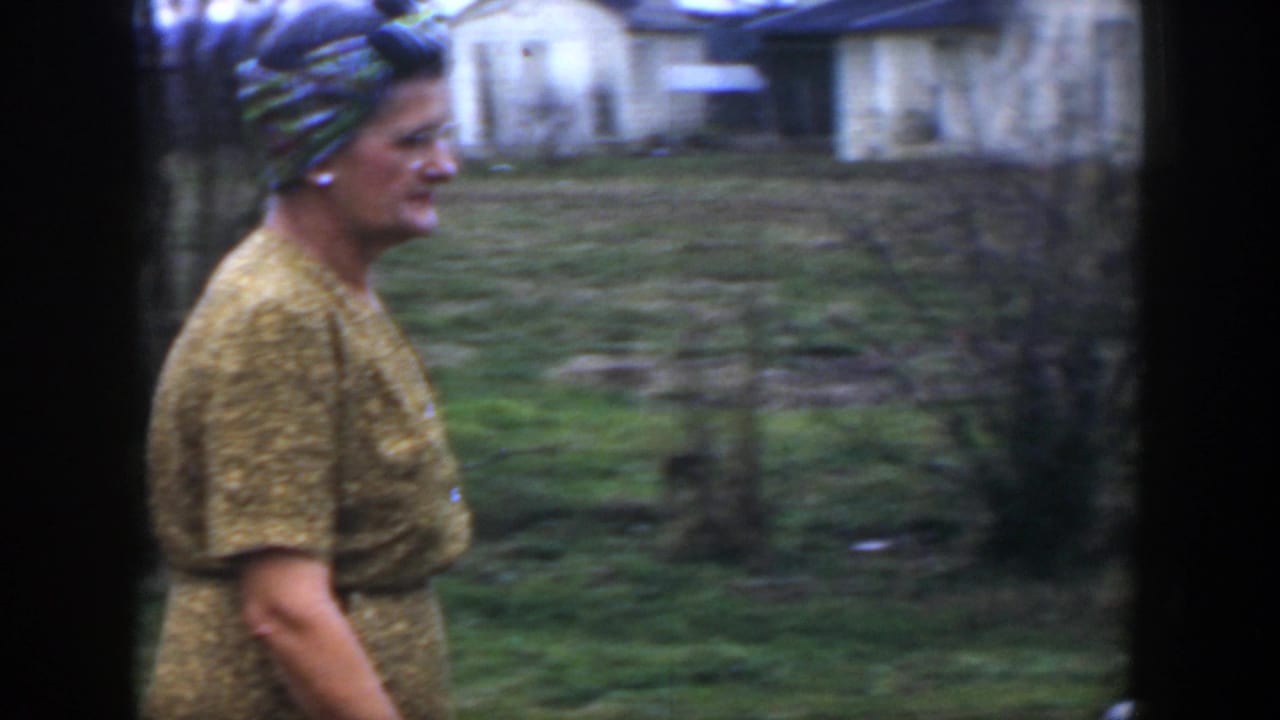 Woman in yellow walks in profile across grass with a house behind her.