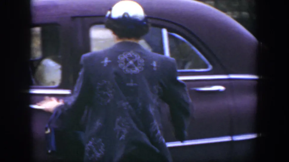 In 1951 Cuba footage, a woman seen from behind in a dark embroidered jacket reaches toward the door area of a purple car.