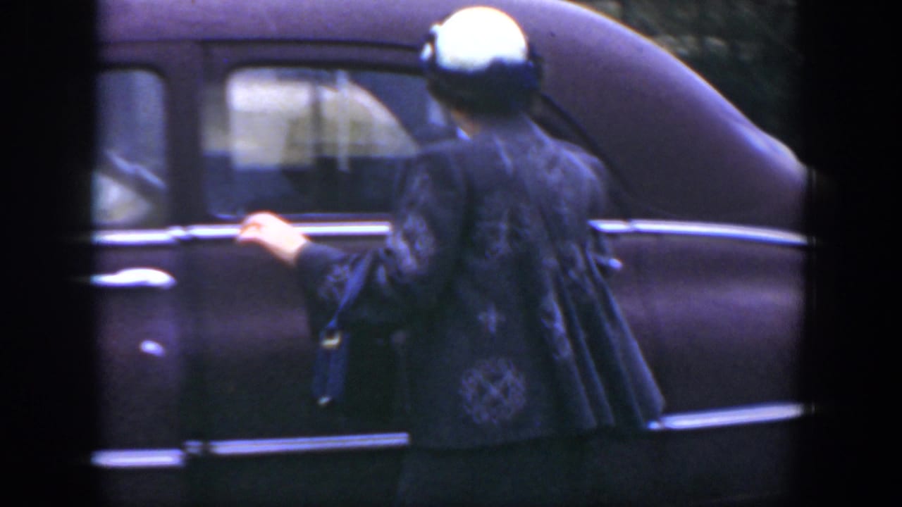 Woman in a white hat holds the door of a dark vintage sedan.