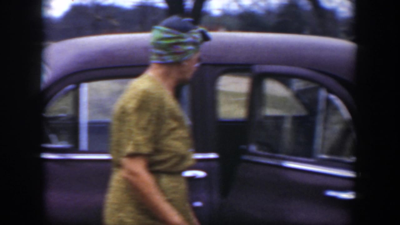 Woman in a patterned dress and headscarf walks beside the sedan.