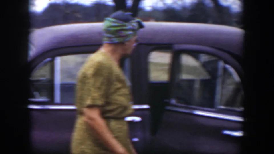 A woman in a patterned dress and colorful headscarf moves alongside the dark sedan in the Cuba 1951 footage.
