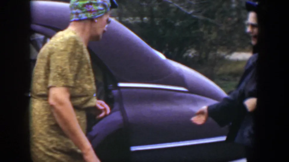 Two adult women stand near the sedan, with one in a patterned dress and another in dark clothing partly visible.