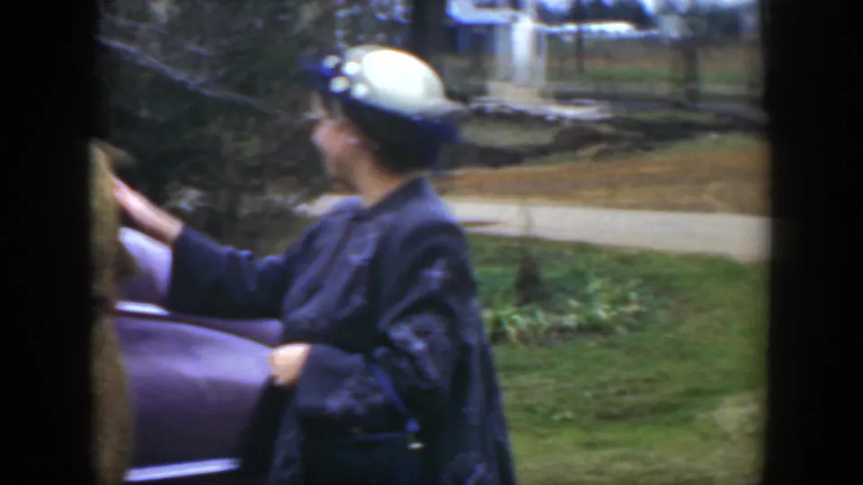 A woman in a white hat and dark embroidered jacket holds the open sedan door in the 1951 Cuba home-movie footage.