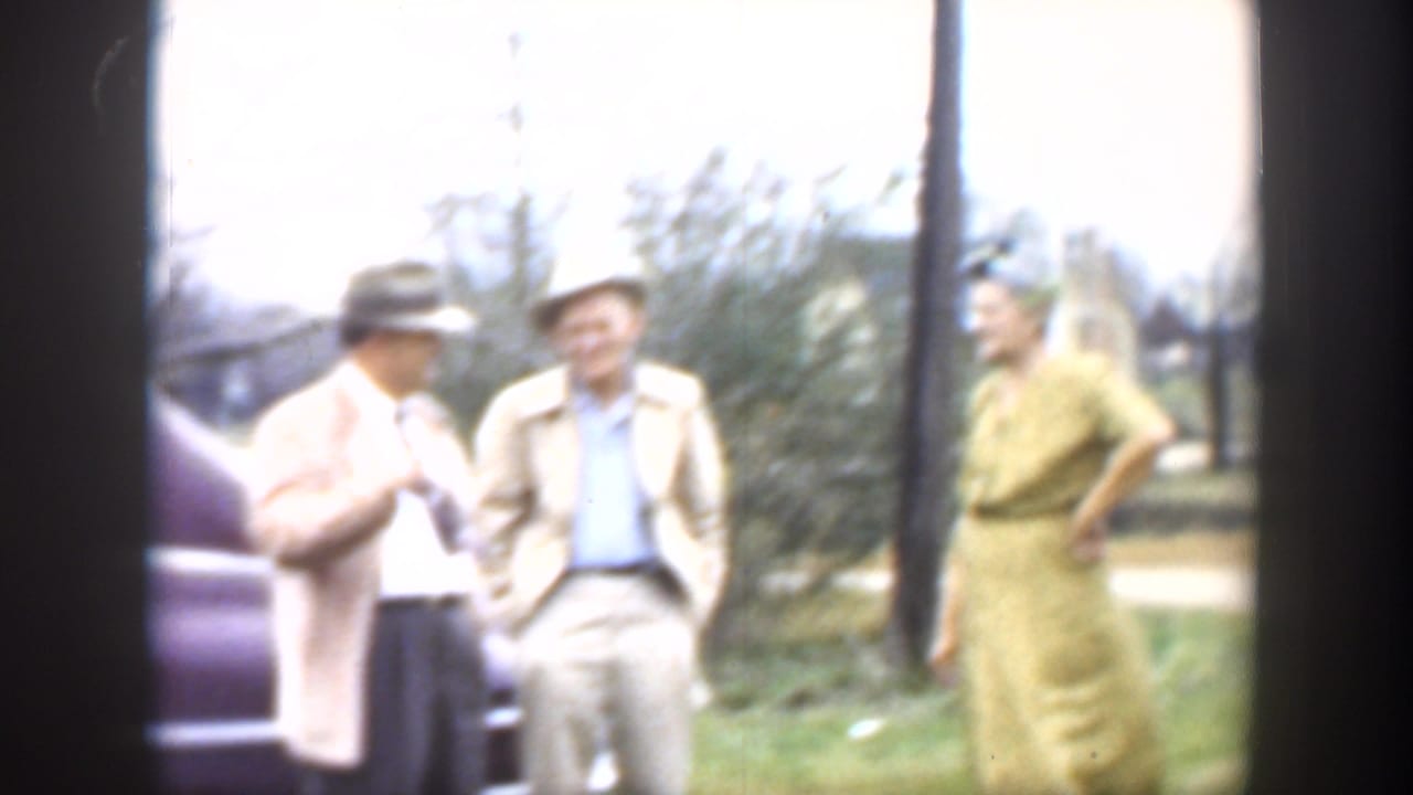 Three adults standing outdoors beside a partial dark car, with trees and utility poles behind them.