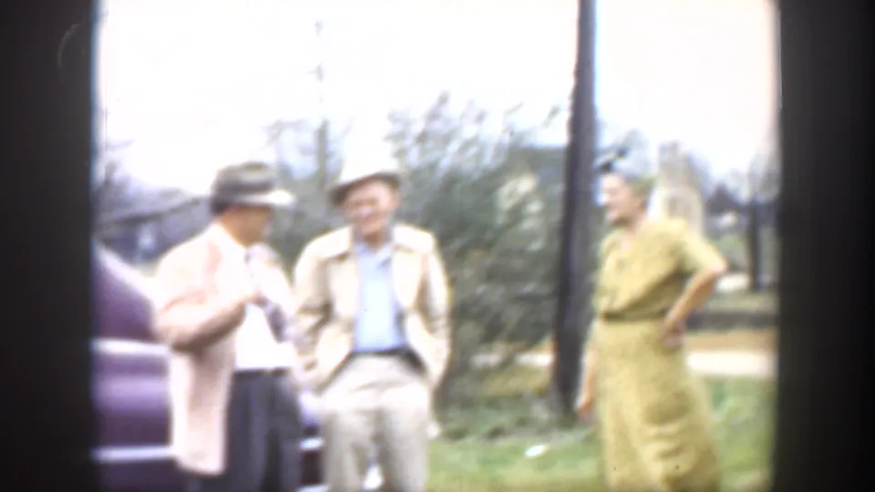1951 Cuba home-movie frame showing three adults in mid-century attire socializing outdoors near a partial dark car.