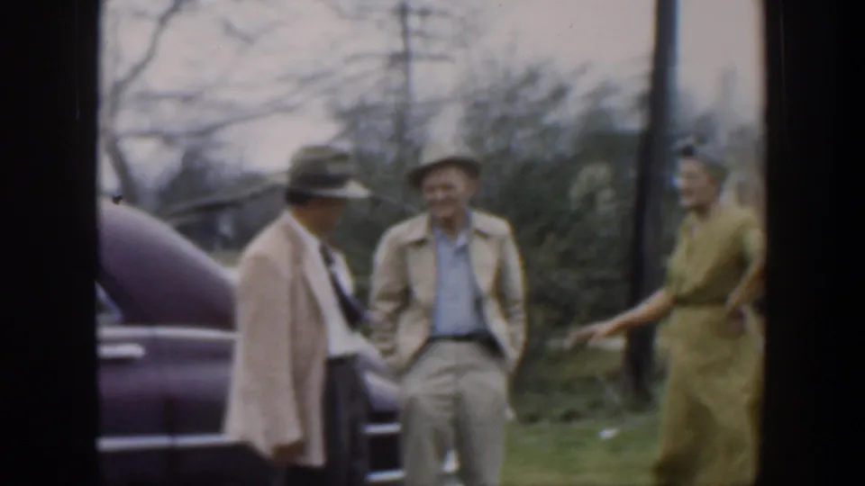 1951 Cuba 8mm frame of two hatted men and a woman in a green dress conversing near a parked car.