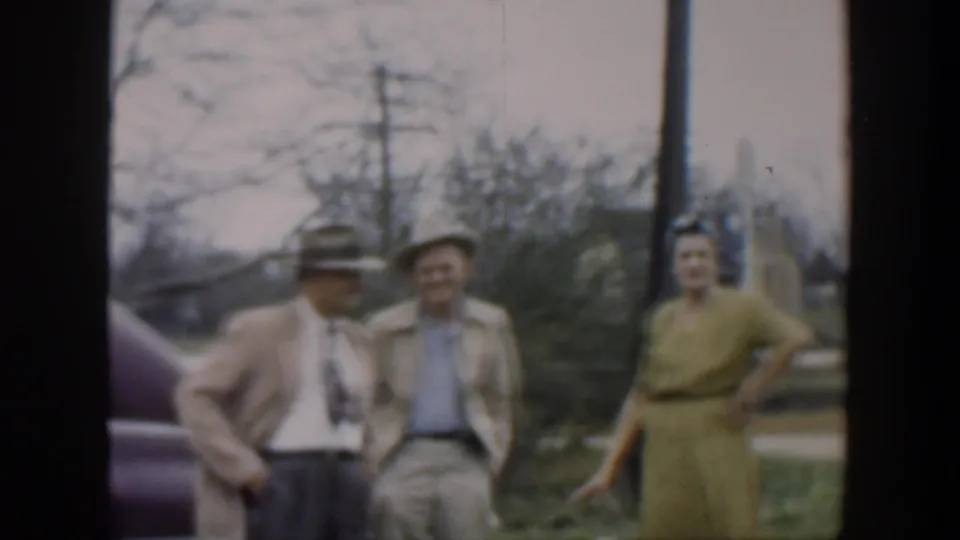 1951 Cuba home-movie frame showing adults in hats and a green dress standing in a yard beside a partial car.