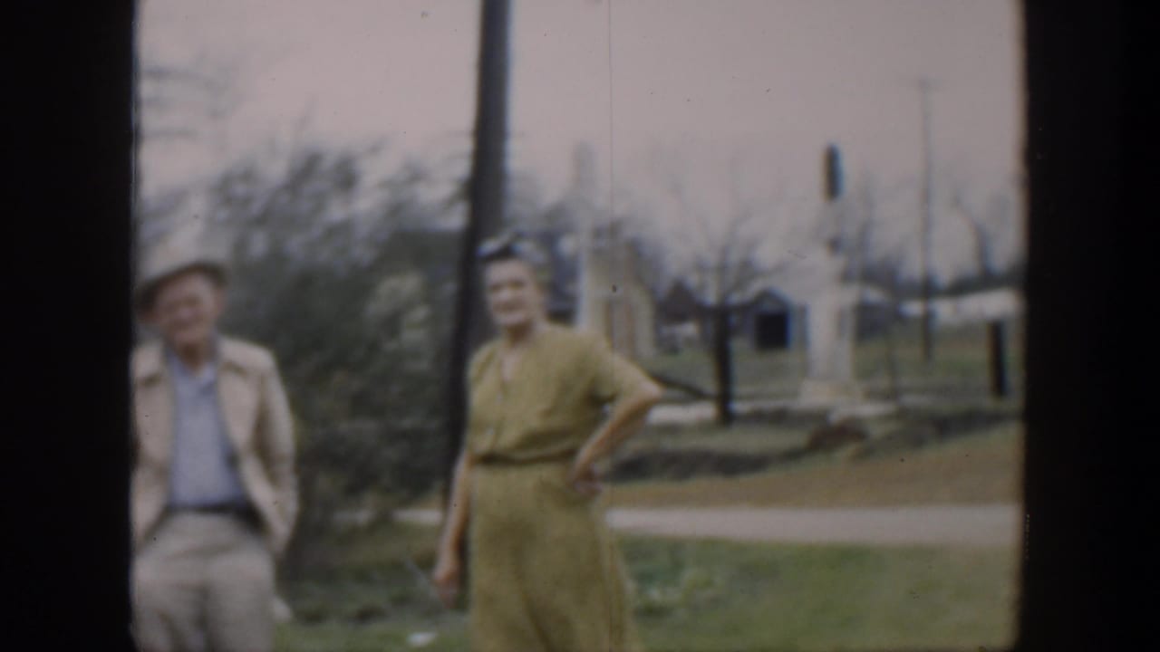 In 1951 Cuba home-movie footage, a woman in a yellow dress and a man in a light jacket stand near a brown pole with trees and utility poles behind them.
