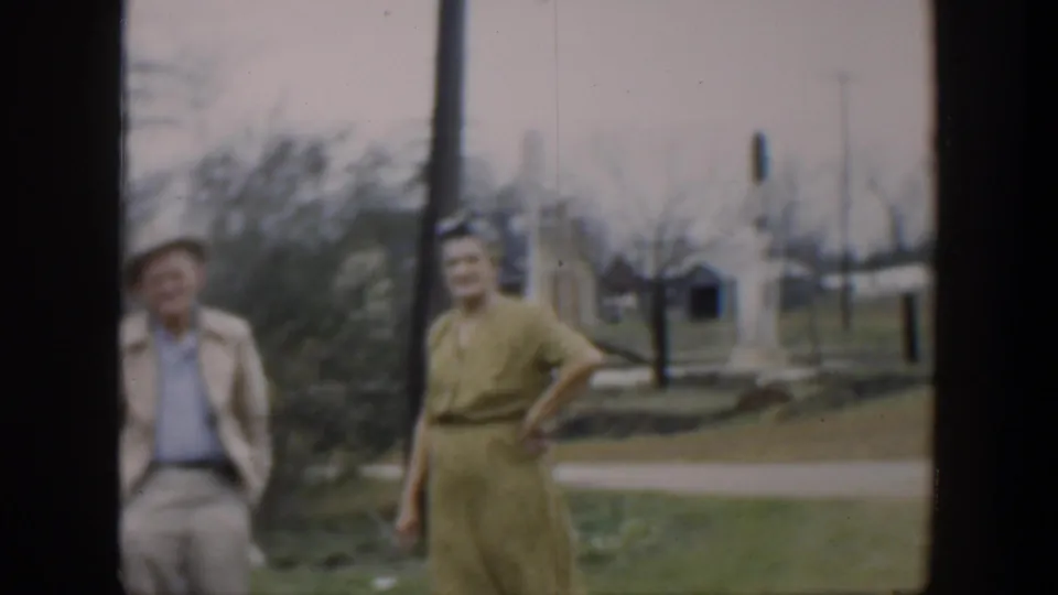 In 1951 Cuba home-movie footage, a woman in a yellow dress and a man in a light jacket stand near a brown pole with trees and utility poles behind them.