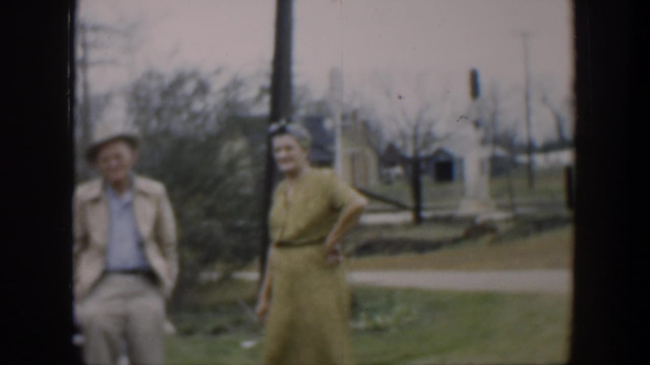 Woman in yellow dress faces the camera while a man in a hat stands beside her.