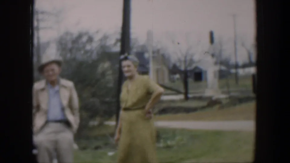 The woman in a yellow dress faces the camera in an outdoor Cuba setting, with a man in a hat, a brown pole, trees, and utility poles visible nearby.