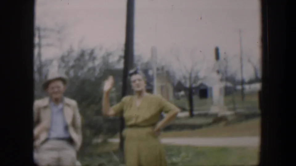 The woman in a yellow dress waves at the camera while the man in a hat remains beside her, with a lawn, roadside, and utility poles in the background.