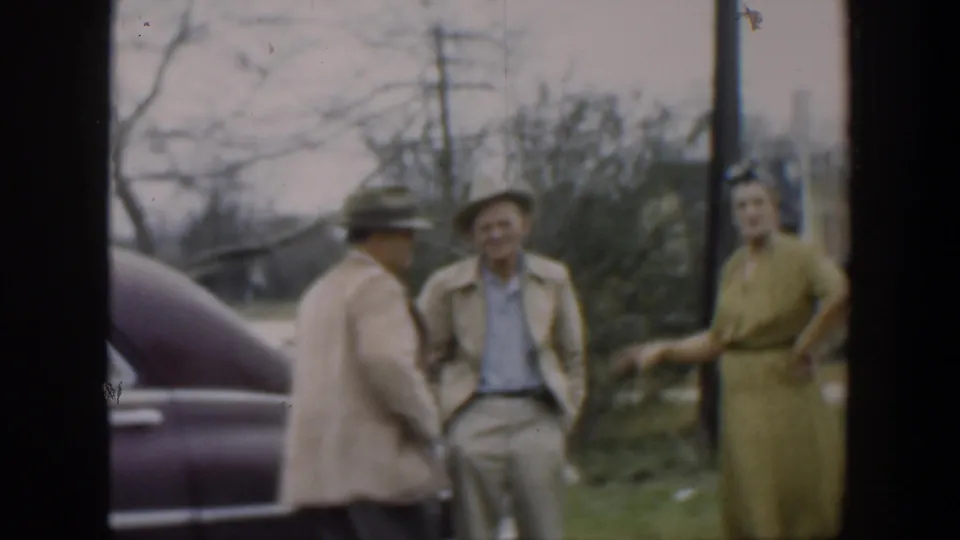 In 1951 Cuba home-movie footage, two men in hats and a woman in a green dress stand beside a purple vintage car outdoors.