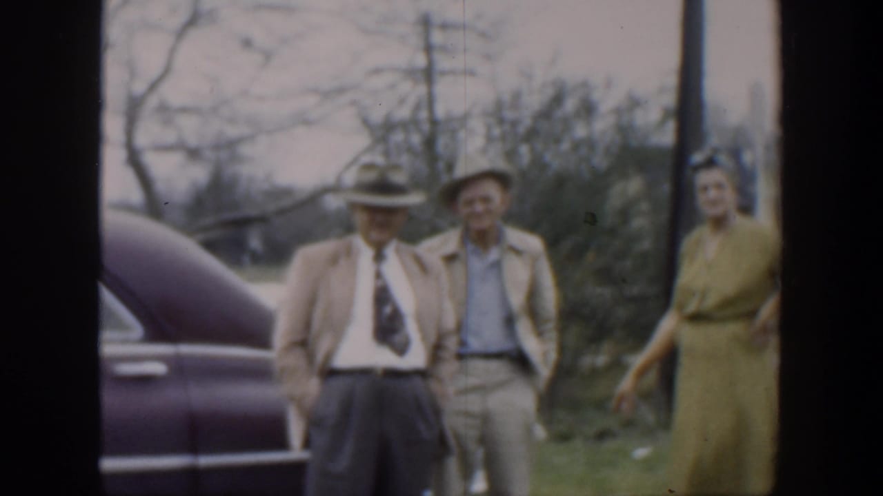 Two men stand by a car while a woman in a green dress appears at the right edge.