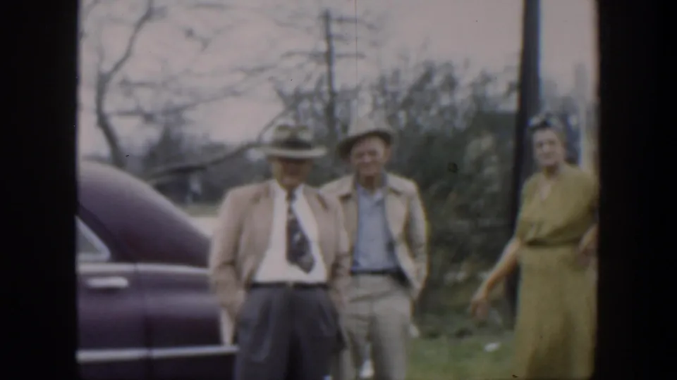 Two well-dressed men in hats stand near a purple car as a woman in a green dress moves at the edge of the 1951 Cuba frame.