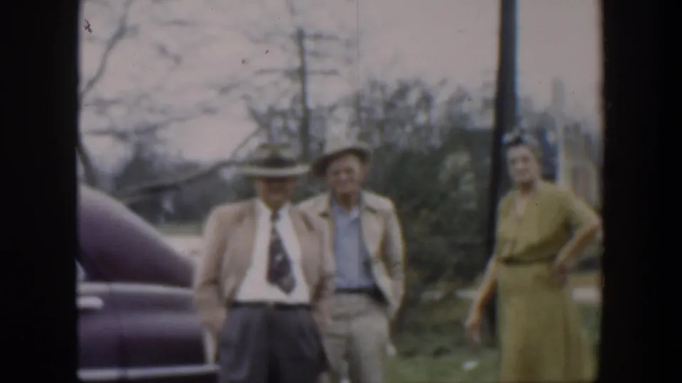 The 1951 Cuba home-movie frame shows two men in hats and a woman in a green dress standing by a parked vintage car.