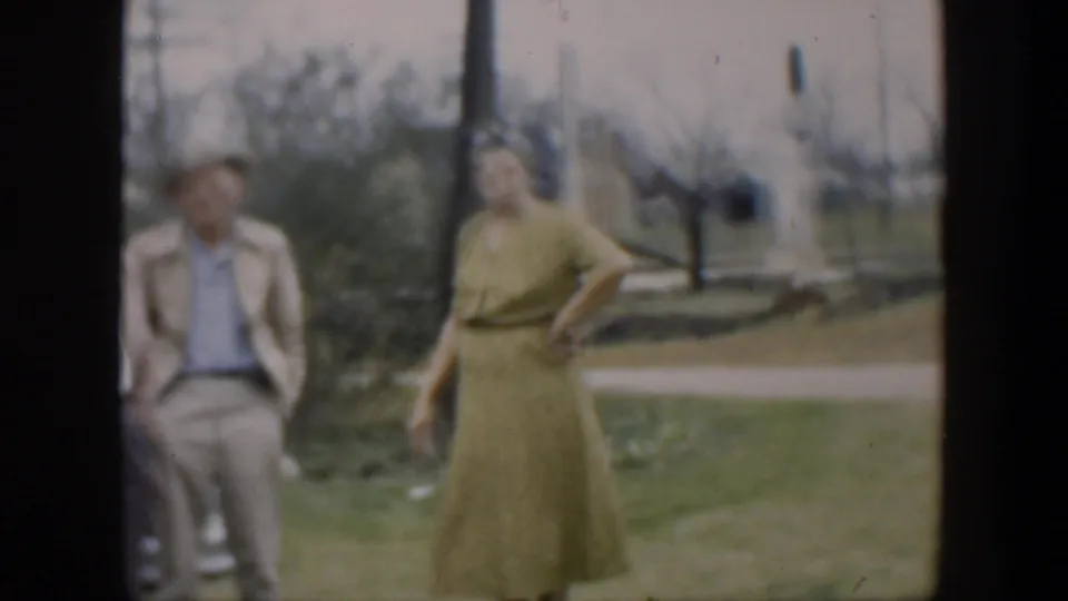 A woman in a green dress stands in the foreground while two men remain near the car, with a light-colored building in the background.