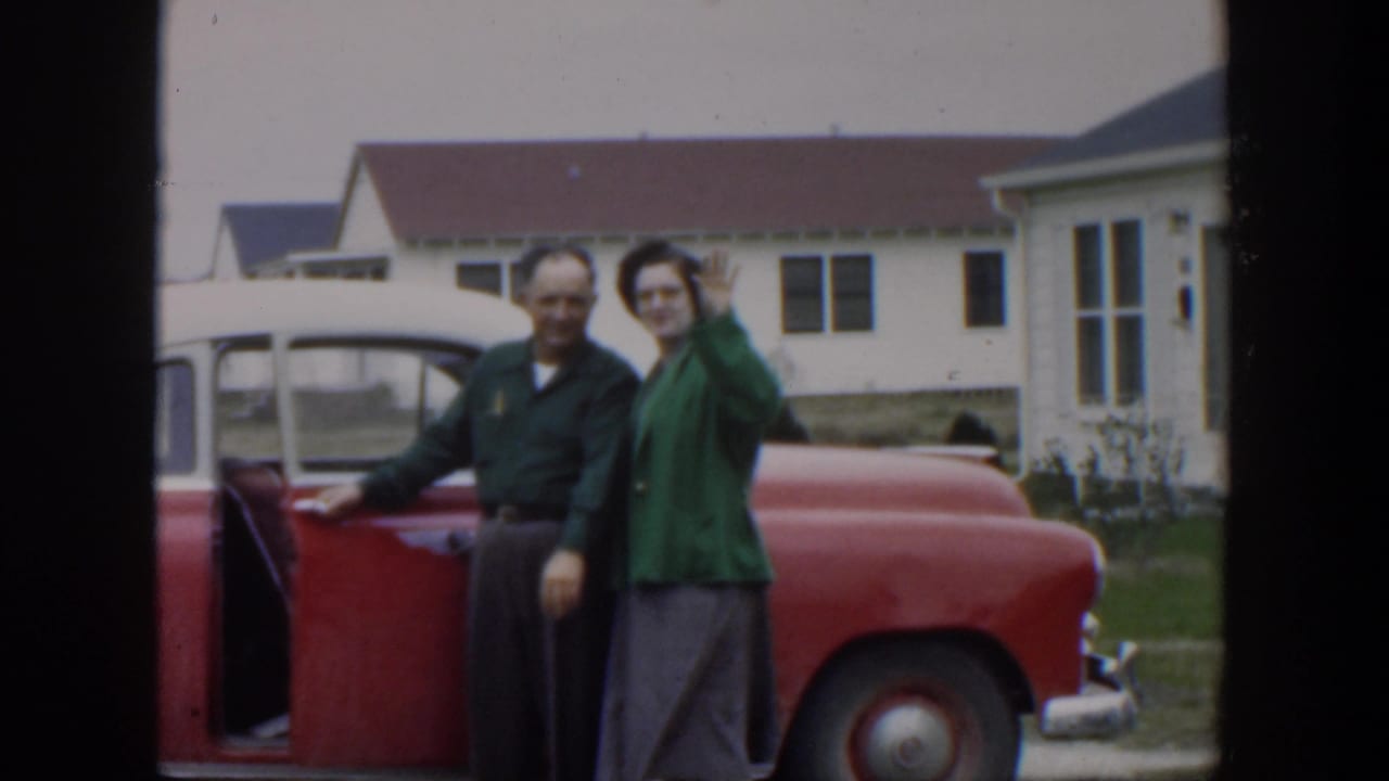 Man and woman in green jackets stand beside a red car and wave near houses.
