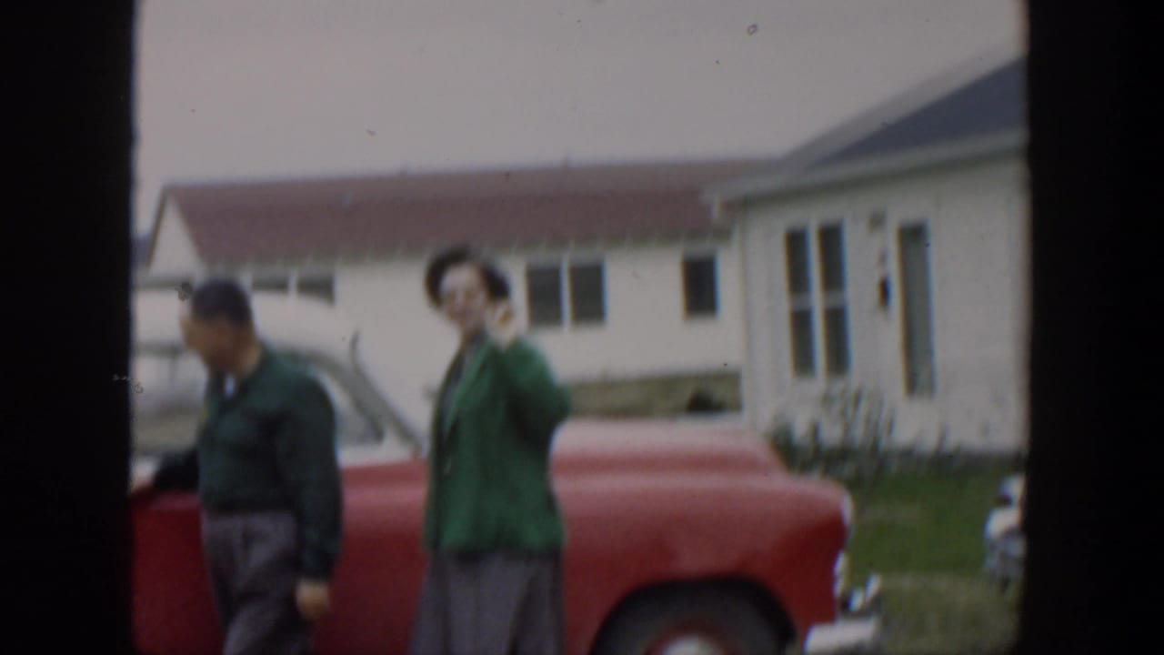 Woman waves beside a red car while the man turns toward the open door.