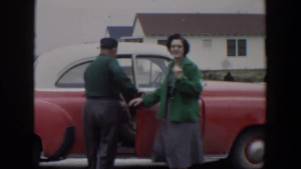 The man handles the door of the red car as the woman faces the camera, continuing the 1951 Cuba home-movie departure.