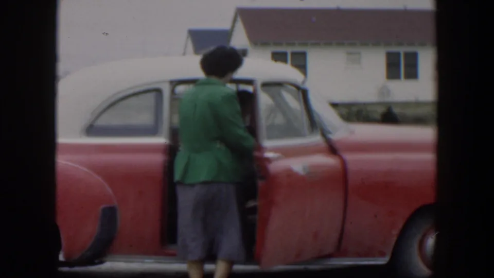 In 1951 Cuba home-movie footage, a woman in a green jacket and dark hat stands by the open door of a red-and-white car.
