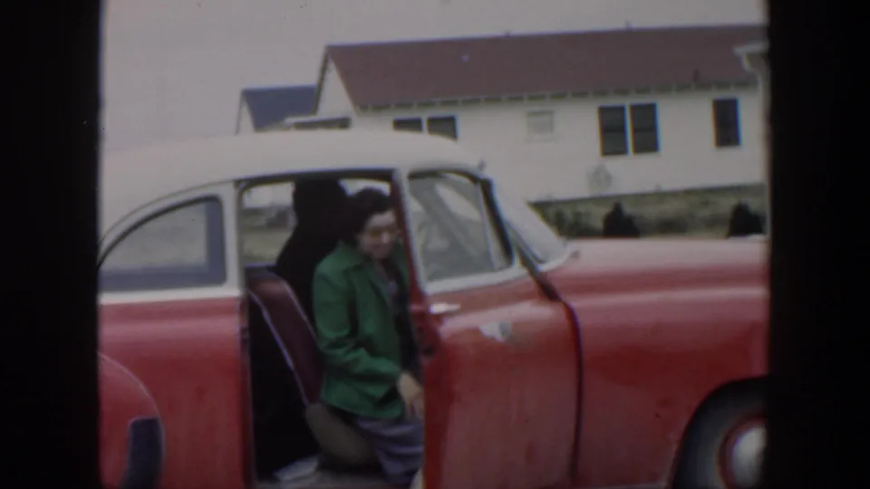 The woman sits into the red-and-white car with the door open, with houses visible behind her in the 1951 Cuba home movie.
