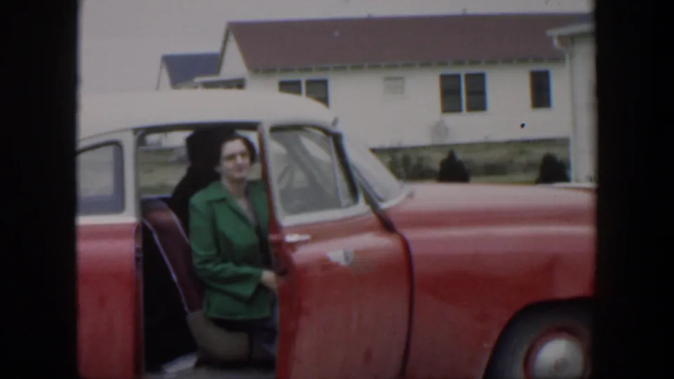 Seated at the open door of the red-and-white car, the woman looks toward the camera in a quiet residential scene.