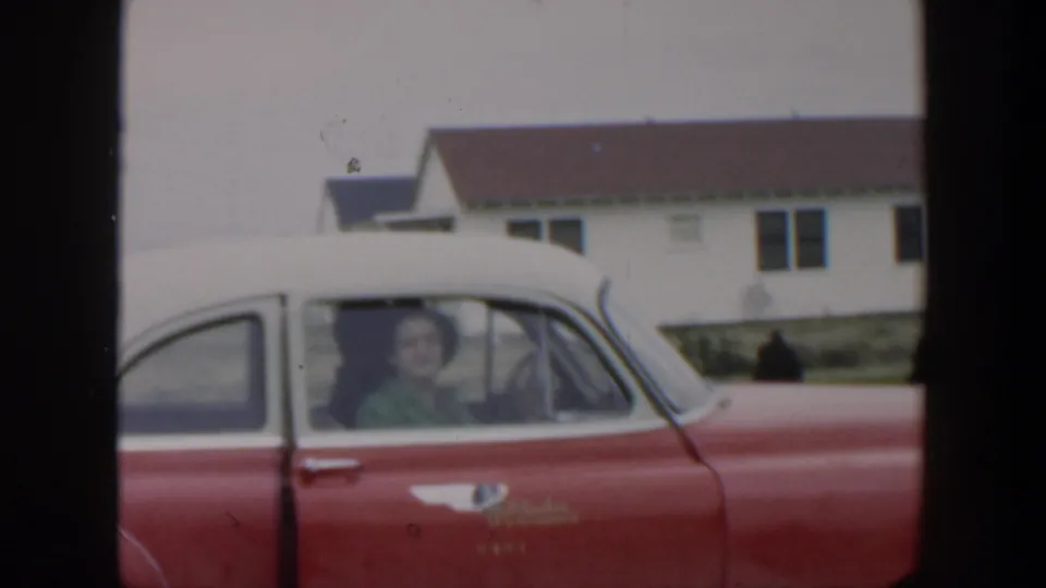 The woman appears inside the red-and-white car behind the side window, with residential buildings in the background.